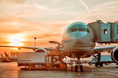 Modern passenger airplane parked to terminal building gate at airside apron of airport with close up airplane parts jet engine wing windows gear tow tractor noon sun view air travel background